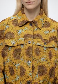 Femme portant une veste jaune à motif tournesol, fermeture à boutons sur le devant et deux poches poitrine à rabat boutonné.