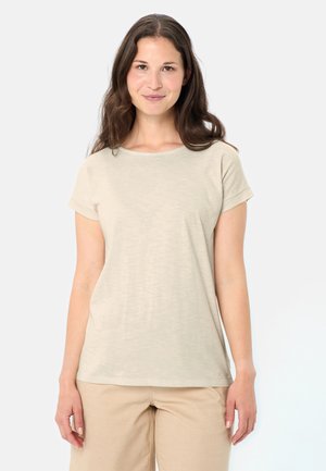 Donna in piedi e sorridente, indossa una maglietta beige a maniche corte e pantaloni color crema chiaro, su uno sfondo bianco uniforme.