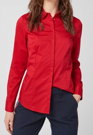 Mujer con una camisa roja ajustada abotonada y pantalones azul marino con una franja lateral, con la mano en el bolsillo, frente a un fondo liso.