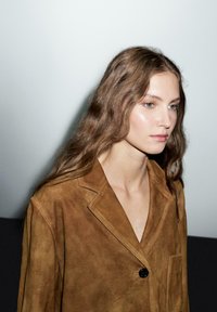 Blazer in suede marrone con colletto a scollo, bottoncino nero singolo e tasca. La modella ha capelli lunghi e mossi e un trucco neutro.