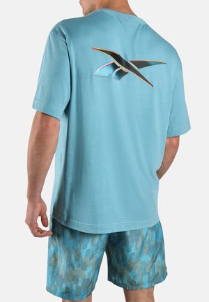 Mann trägt ein hellblaues T-Shirt mit einem schwarzen und orangefarbenen abstrakten Logo auf dem Rücken und blaue Shorts mit Camouflage-Muster, steht mit dem Rücken zur Kamera.