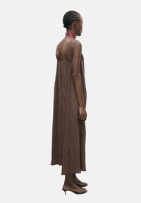 Rochie maxi maro, fără mâneci, cu o croială lejeră, realizată dintr-un material texturat. Are un decolteu adânc la spate și bretele subțiri, asortată cu sandale plate.