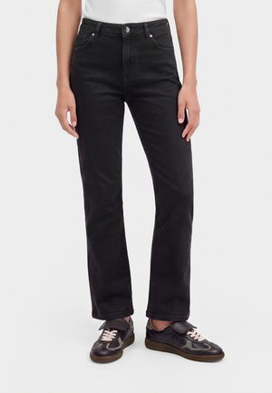 Persoon die zwarte jeans met hoge taille en rechte pijpen draagt en zwarte sneakers met roze veters tegen een effen witte achtergrond.