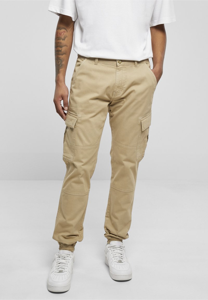 Hombre con pantalones cargo beige con puños elásticos y zapatillas blancas, de pie con las manos en los bolsillos sobre un fondo liso.