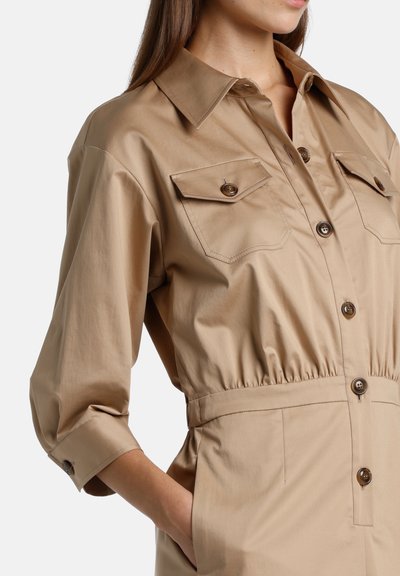 Beige shirt dress with a collar, button closure, and two chest pockets. Features elastic waist and cuffed sleeves for a relaxed fit.