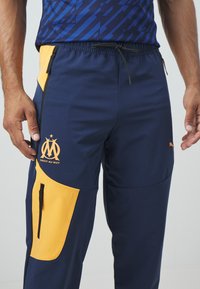 Marineblauwe sportbroek met gele accenten, voorzien van een logo en ritszakken. Gemaakt van gladde stof met een elastische tailleband en een trekkoord.