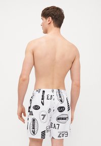 Pantaloni da bagno bianchi con motivi grafici e loghi neri, con vita elastica e vestibilità ampia, mostrati da dietro.