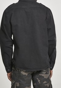 Svart bomullsskjorta med lång ärm och knäppt manschett. Har en ryggsömn och rundad nederkant. Matchas med kamouflagebyxor.