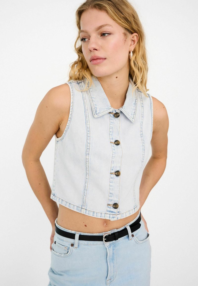 Τζιν αμάνικο crop top με γιακά, ανοιχτό μπλε χρώμα, πέντε καφέ κουμπιά μπροστά και έντονες ραφές.