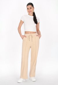Beige elastiske bukser med taljeelastik og sidelommer, kontrasterende syninger, parret med en hvid cropped top og hvide sneakers. Glat stoftekstur.