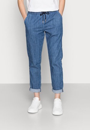 Person iført op-rullede blå bukser med snor, hvide sneakers og en hvid skjorte, stående med hænderne i lommerne.