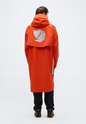 Personne portant un long manteau orange avec capuche, logo de loup réfléchissant dans le dos, pantalon noir et chaussures de plein air, vue de dos.