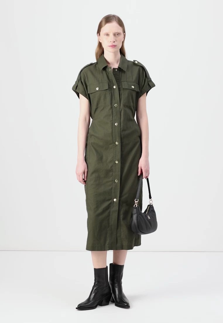 Robe vert olive à manches courtes et fermeture boutonnée, avec deux poches poitrine et une silhouette droite ; associée à des bottines noires et un petit sac à main.