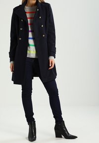 Abrigo largo navy con botones dorados, combinado con un suéter gris a rayas con franjas multicolores, jeans oscuros y botas negras de tobillo.