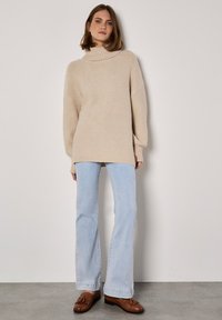 Pull beige oversize avec un col roulé haut, des manches longues et des fentes sur les côtés, associé à un jean bleu clair évasé et des mocassins marron.
