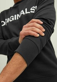 Schwarzer Sweatshirt aus Baumwolle mit weißem Text "ORIGINALS" auf der Brust. Rippbündchen an den Ärmeln und am Saum sorgen für Textur.