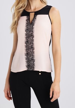 Top beige sin mangas con una franja vertical de encaje negro en el centro y escote negro, combinado con pantalones negros.