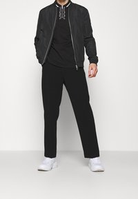 Černá bomber bunda na zip, černé tričko s bílým grafickým designem, volné černé kalhoty a bílé tenisky.