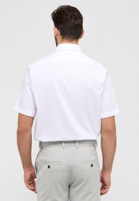 Camicia bianca a maniche corte con colletto tradizionale, texture liscia e design dritto sul retro, abbinata a pantaloni grigi chiari con tasche.