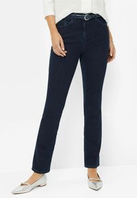 Jean droit en denim foncé avec taille mi-haute, design à cinq poches et une ceinture avec boucle argentée. Associé avec des ballerines argentées.