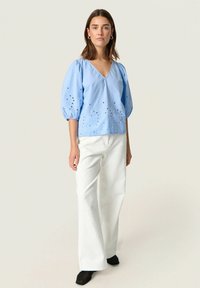 Top in cotone azzurro chiaro con maniche a sbuffo, scollatura a V, caratterizzato da dettagli ricamati a occhio di pernice. Abbinato a pantaloni bianchi larghi e strutturati.
