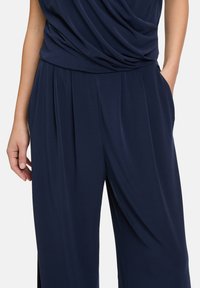 Navy blue jumpsuit with draped top, pleated front, and side pockets. Made of soft fabric, featuring a relaxed fit and smooth texture.