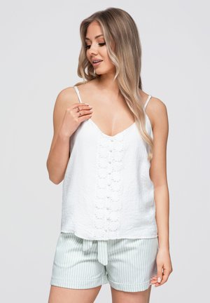 Vrouw met lang blond haar die een witte mouwloze top met bloemendetails en lichtgroene gestreepte shorts draagt, kijkt naar beneden en raakt een bandje aan.