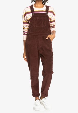 Salopette en velours côtelé marron avec bretelles réglables et une poche frontale, associée à un t-shirt à manches longues rayé et des baskets blanches.