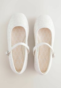 White ballet flats with glittery upper, rounded toe, and a strap across the instep. Interior features a light pattern and scalloped edging.