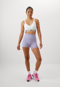 Hvit sportbralett med svart Nike-logo bånd, kombinert med lavendel shorts. Modellen har på seg rosa joggesko med hvite sokker. Enkel, tettsittende design.