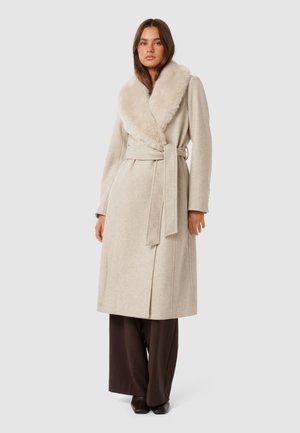 Beige Wollwickelmantel mit einem breiten, weichen Kunstfellkragen, langen Ärmeln und einem Bindegürtel. Kombiniert mit dunkelbraunen weit geschnittenen Hosen.