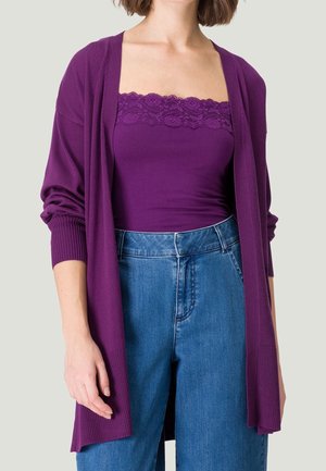 Femme portant un caraco violet orné de dentelle, un cardigan assorti à ouverture devant, et un jean taille haute bleu sur un fond neutre.