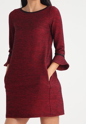 Vrouw draagt een rode jurk met textuur tot op de knie met driekwartklokmouwen en zakken, staand tegen een effen lichte achtergrond.