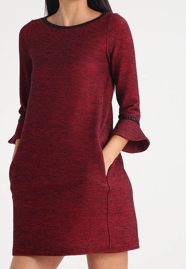 Mujer con un vestido rojo texturizado hasta la rodilla, con mangas acampanadas de tres cuartos y bolsillos, de pie frente a un fondo liso y claro.