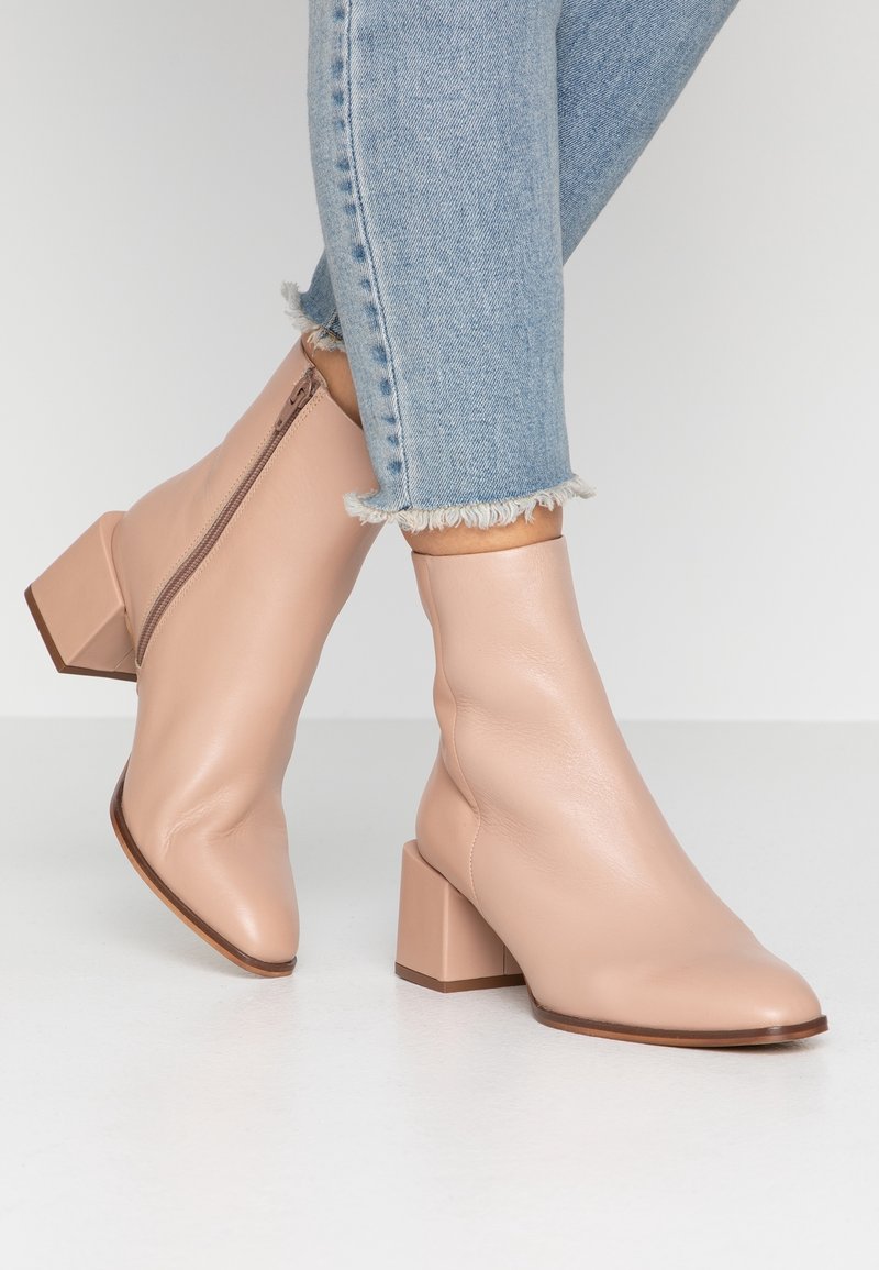 Femme portant des bottines en cuir rose pâle avec des talons carrés et des zips sur le côté, associées à un jean bleu clair à ourlet effiloché.