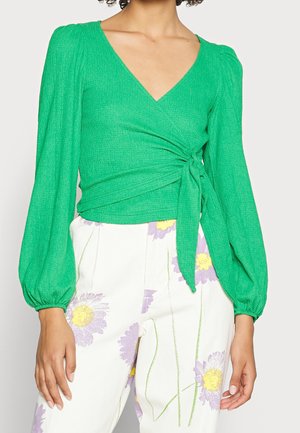 Mujer con un top cruzado de manga larga de color verde brillante con un lazo lateral y pantalones blancos con estampados florales en púrpura y amarillo.