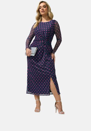 Vrouw draagt een marineblauwe midi-jurk met roze polkadots, doorschijnende lange mouwen, een zijsplit, een zilveren clutch en zilveren sandalen met hoge hak.