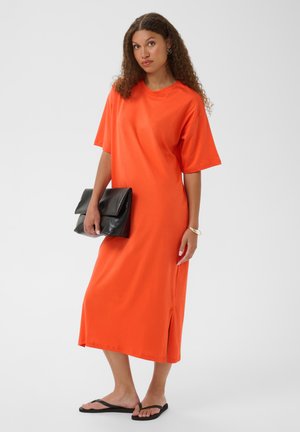 Femme aux cheveux bouclés portant une robe midi orange, des tongs noires, tenant une pochette noire, debout sur un fond uni et clair.