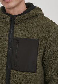 Olijfgroene fleecejack met een zwarte stoffen ritszak, gestructureerde buitenkant, volledige rits aan de voorkant en met capuchon.