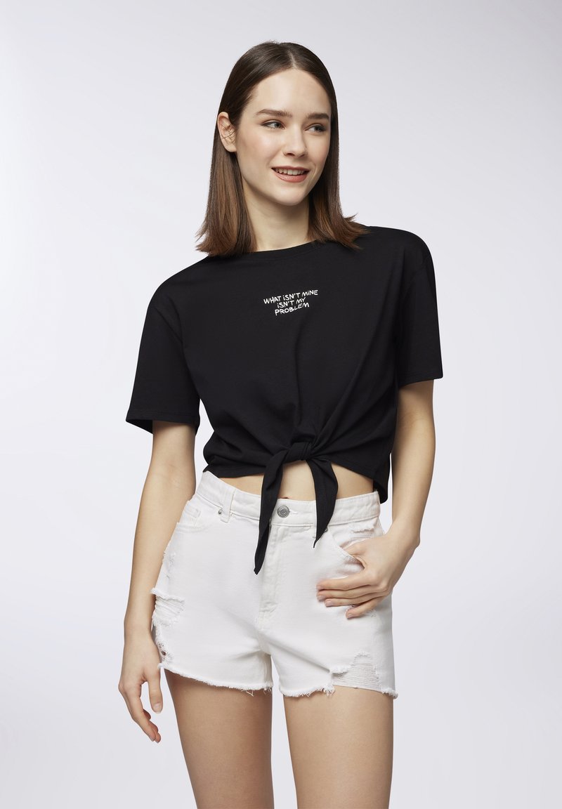 T-shirt nera corta con dettaglio del nodo frontale, caratterizzata da testo bianco, abbinata a pantaloni corti in denim bianco strappati. Texture del tessuto liscia.