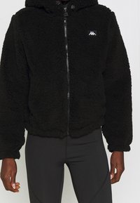 Felpa nera con zip realizzata in un tessuto fluffy e testurizzato. Presenta un cappuccio, polsini elasticizzati e un piccolo logo sul petto.