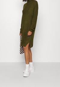 Person som bär en olivgrön stickad långärmad klänning med sidofickor, vita sneakers, vita strumpor och håller en svart-vitt rutig väska.