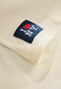 Kreemi värvi kangas, mille servas on tumehall etikett, kus on valge ja punase tekstiga "I ♥ NY".