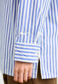 Striped skjorte med blå og hvite vertikale striper, bomullsmateriale, knappede mansjetter og en hvit logo på ermet.
