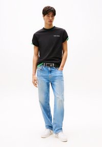 Sort T-shirt med grønne accenter, med "TOMMY JEANS" logo. Lyst blå slidte jeans, parret med hvide sneakers. Afslappet pasform.