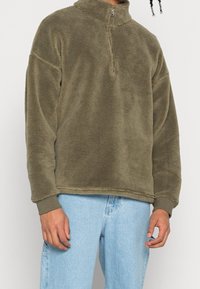 Maglione in pile verde oliva con collo a mezza zip, vestibilità ampia, spalle scese e polsini a costine; tessuto morbido e design casual.