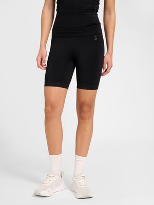 Person, die schwarze enganliegende Shorts, ein schwarzes Oberteil, weiße Socken und weiße Sportschuhe trägt, steht vor einem grauen Hintergrund.