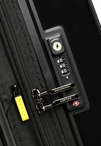 Valigia nera con superficie testurizzata, dotata di una serratura approvata TSA, doppie cerniere argentate e un'etichetta gialla Delsey.