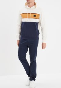 Sudadera con capucha con bloques de color crema, mostaza y azul marino; bolsillo tipo canguro; tela de mezcla de algodón; combinada con pantalones azul marino y zapatillas blancas.