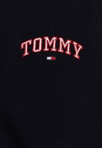 Čierny pletený materiál s vyšívaným nápisom "TOMMY" červenou farbou, s malou akcentaciou v červenej, bielej a modrej farbe pod písmenami.